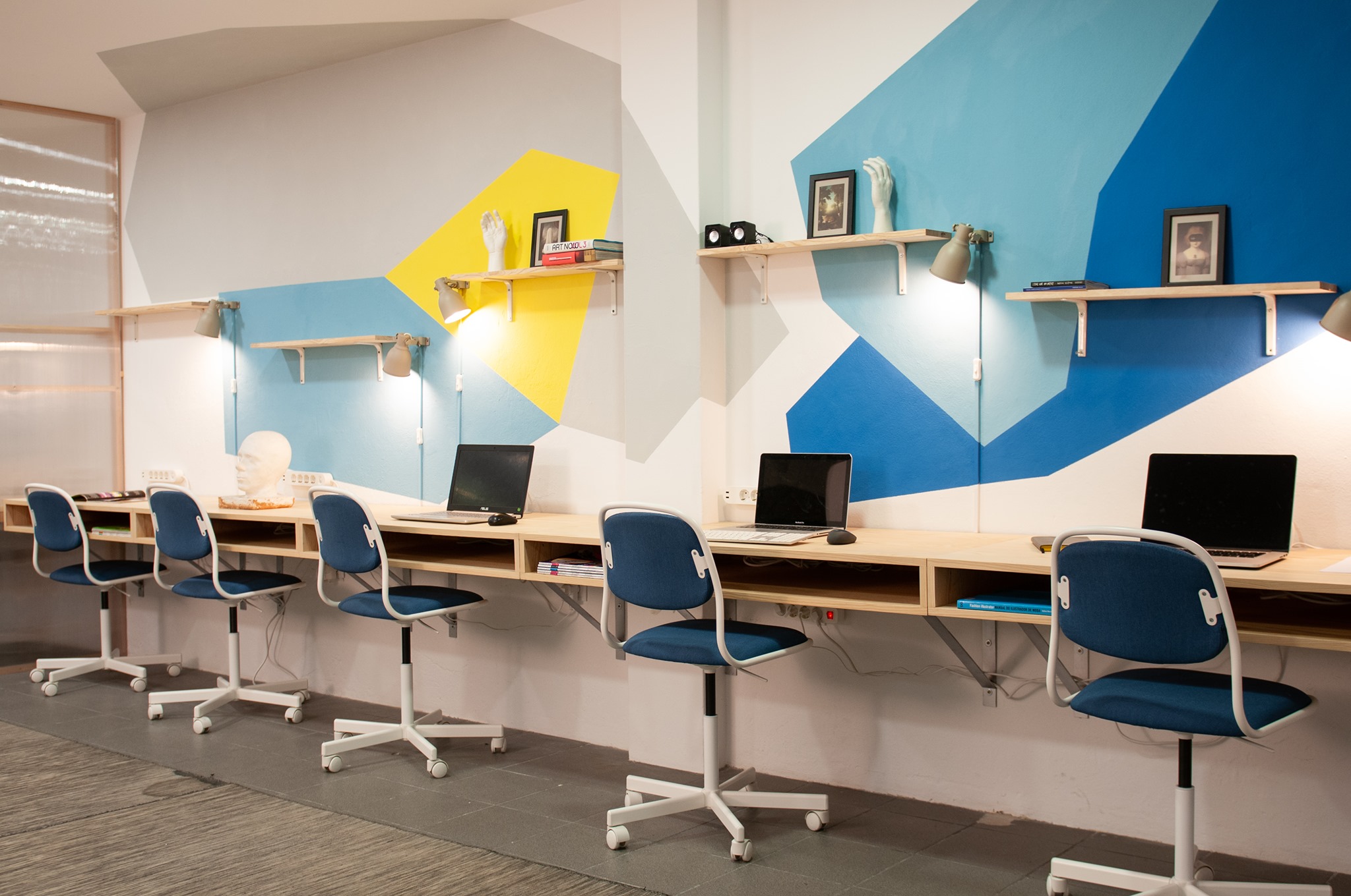 zdjęcie przedstawia salę z rzędem biurek, na kt&oacute;rych stoją laptopy oraz przypisane do nich krzesła biurowe. Na ścianie widać r&oacute;żnokolorowe wzory, w odcieniach niebieskiego i ż&oacute;łci, a także przytwierdzone do ściany p&oacute;łki.