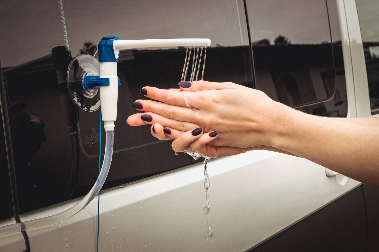Zbliżenie na kobiece dłonie oraz przyczepiony do białej karoserii za pomocą przyssawek kran, z kt&oacute;rego leci woda na kobiece dłonie