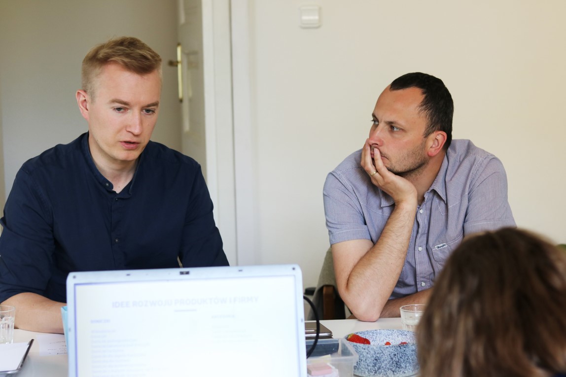 zbliżenie na dwóch młodych mężczyzn siedzących przy stole. Mężczyzna widoczny po lewej stronie patrzy przed siebie, natomiast drugi z mężczyzn patrzy w jego kierunku. Na stole widać komputer typu laptop z otwartym ekranem