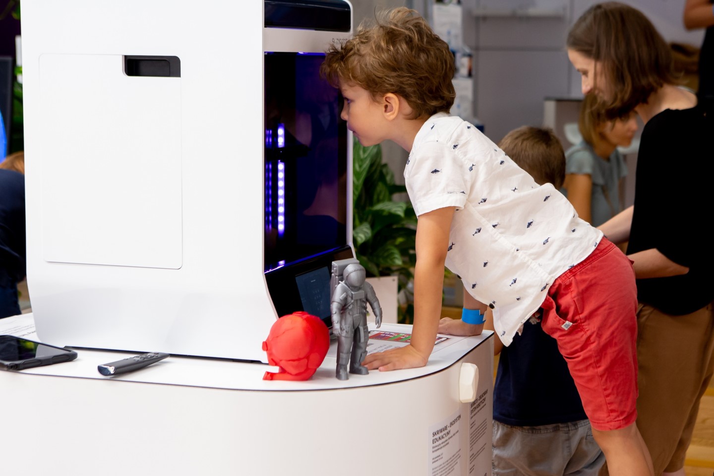 Chłopiec zagląda przez szybę do wnętrza obudowy drukarki 3D. Obudowa drukarki w białym kolorze. Jej szklany front tworzą ciemne szyby. W dolnej części znajduje się panel sterowania drukarki z wyświetlaczem LCD. Obok drukarki stoją dwa wydrukowane modele 3D w niewielkim rozmiarze &ndash; model anatomiczny serca w kolorze czerwonym oraz figurka astronauty w kolorze srebrnym. W głębi zdjęcia widać innego chłopca i dorosłą kobietę.