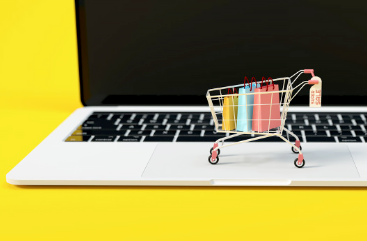 Na zdjęciu widać laptopa na ż&oacute;łtym tle. Na laptopie stoi miniaturowy koszyk zakupowy z trzema malutkimi torbami w środku.