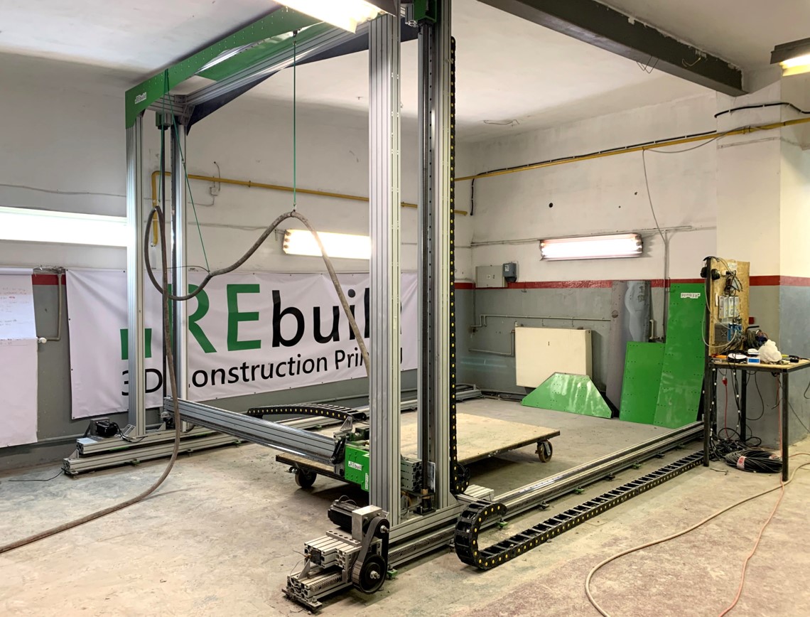 Zdjęcie przedstawia hale przemysłową, w kt&oacute;rej znajduje się metalowa konstrukcja, jej rama jest przytwierdzona do sufitu oraz ściany. Obok leżących na podłodze ramion ramy widać gąsienicę. W środku ramy widać zaś drewnianą płytę, do kt&oacute;rej od spodu przytwierdzone są koła. 