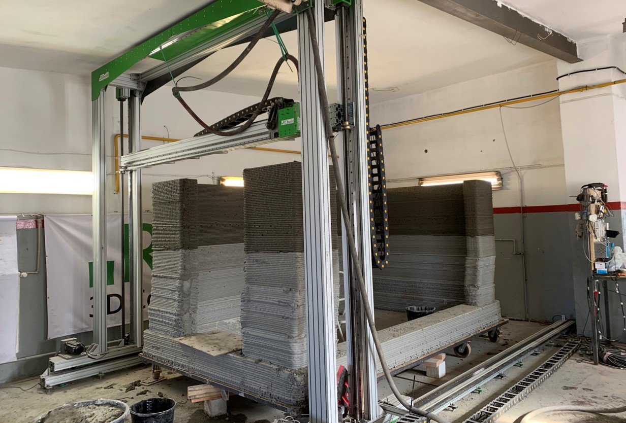 zdjęcie przedstawia hale przemysłową, w kt&oacute;rej znajduje się metalowa konstrukcja, jej rama jest przytwierdzona do sufitu oraz ściany. Obok leżących na podłodze ramion ramy widać gąsienicę. W środku ramy widać zaś drewnianą płytę, do kt&oacute;rej od spodu przytwierdzone są koła. Na drewnianej płycie znajdują się warstwy betonu tworzące ścianę.