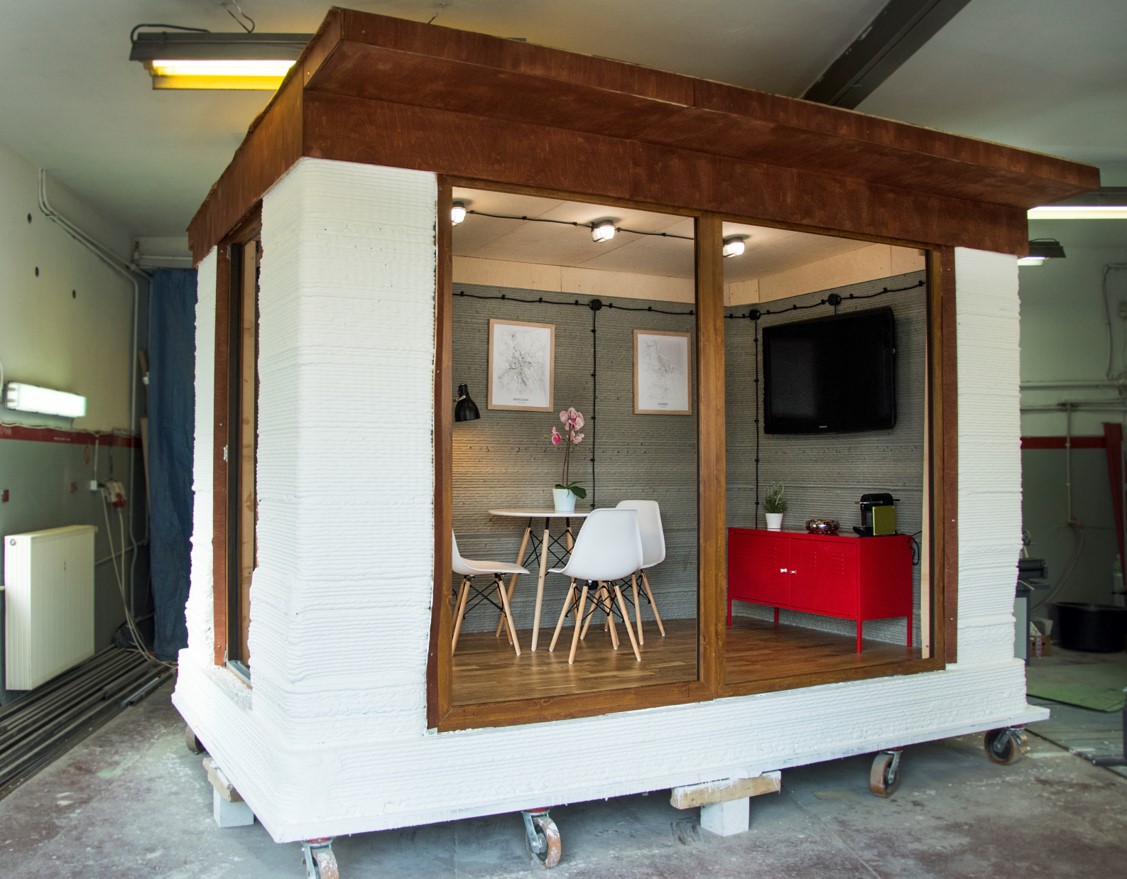 Widok na niewielkich wymiar&oacute;w betonowy domek z centralnie umieszczonym oknem sięgającym od sufitu do podłogi. W środku widać st&oacute;ł, krzesła, komodę oraz wiszące na ścianie telewizor i obrazki.