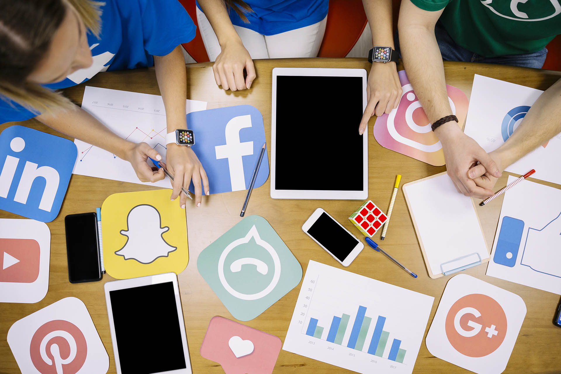 widok od g&oacute;ry na st&oacute;ł, przy kt&oacute;rym siedzi tr&oacute;jka młodych ludzi, dwie kobiety i jeden mężczyzna. Na stole leżą tablety, kartki oraz logotypy ze znakami 