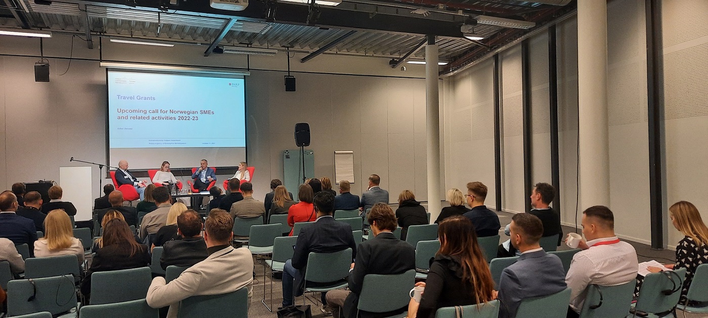 widok na ludzi zgromadzonych na sali konferencyjnej i siedzących na krzesłach na przeciwko sceny, na kt&oacute;rej siedzą cztery osoby, jedna z nich ma mikrofon w ręku