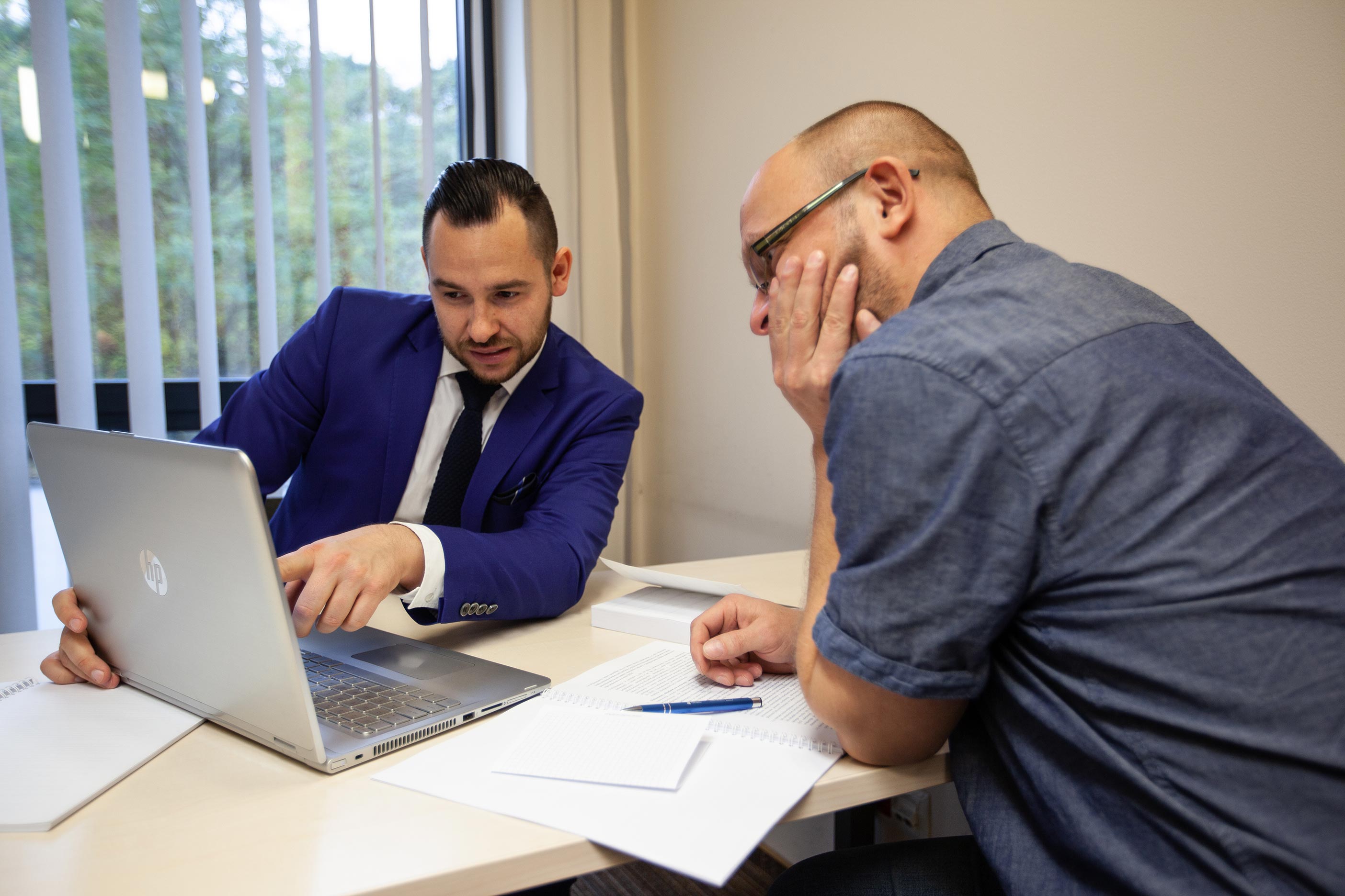 Widok dw&oacute;ch mężczyzn przy biurku, jeden w garniturze pokazuje coś ręką na ekran komputera