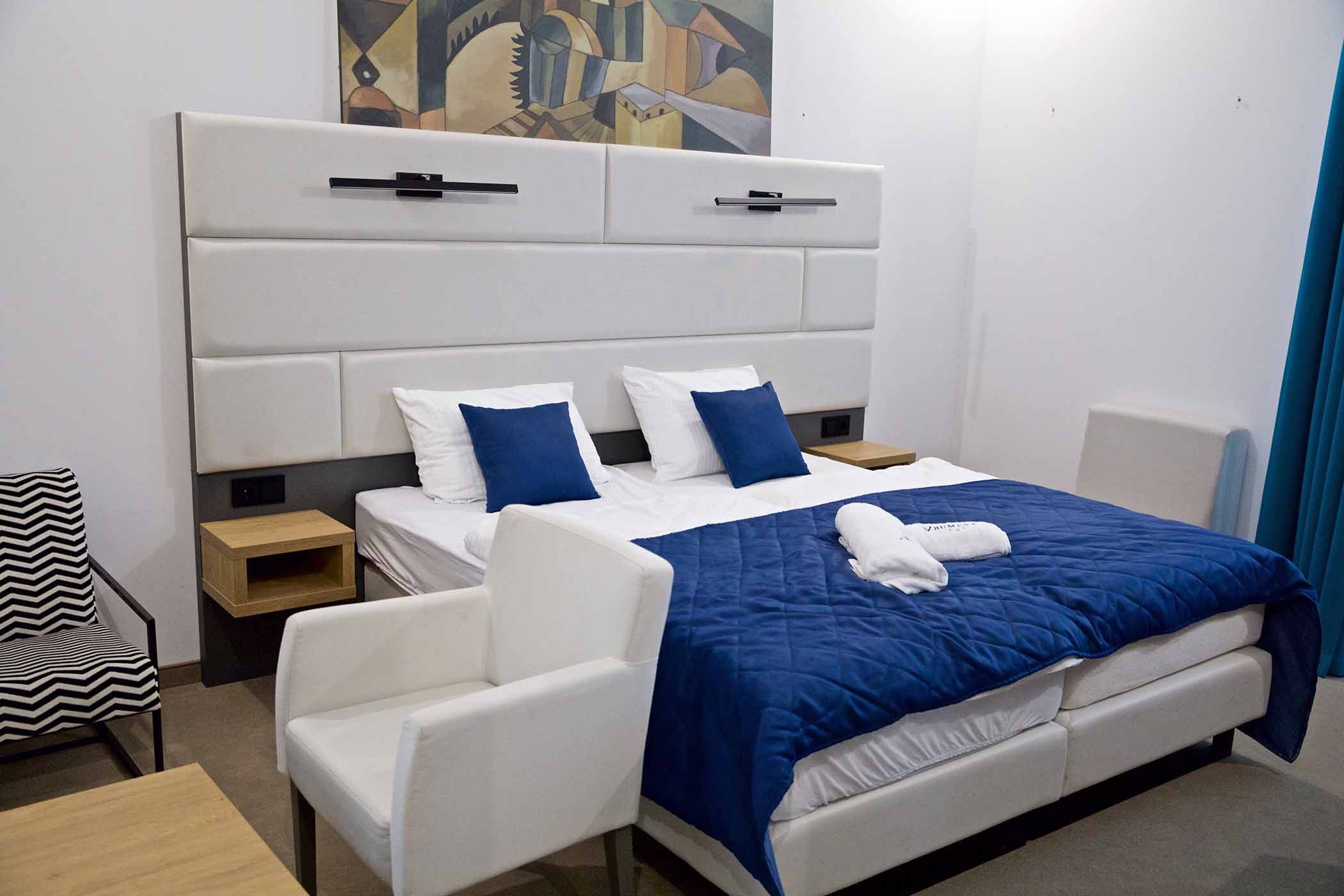 Widok pokoju z meblami jak granatowe krzesło, ł&oacute;żko z białym zagł&oacute;wkiem, drzwi ze zdobioną ościeżnicą