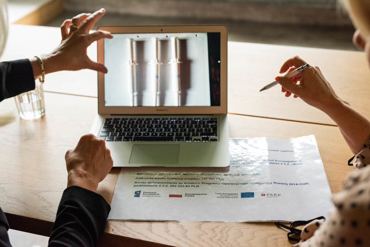 Laptop stoi na biurku, wok&oacute;ł ręce, kt&oacute;re wskazują na ekran.