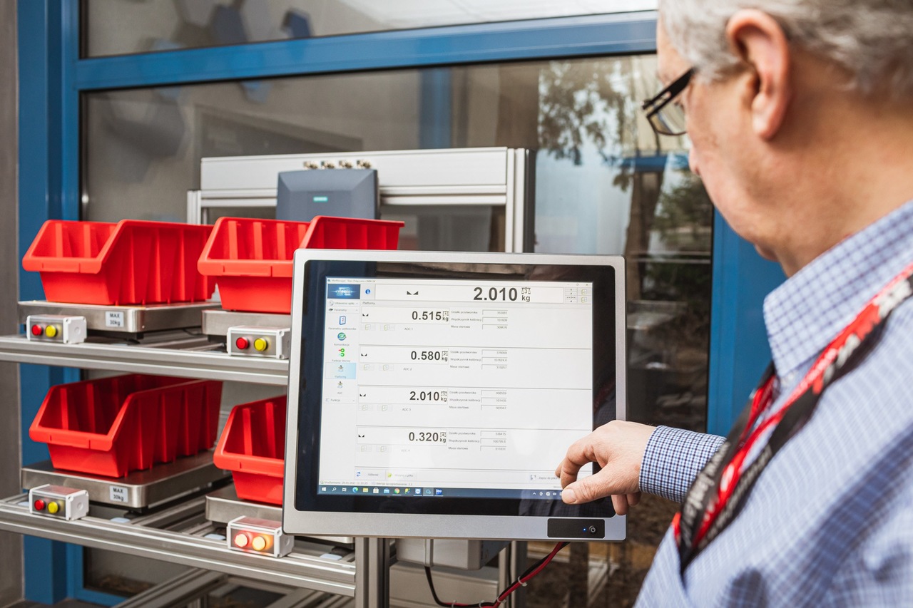 Pojemniki stojące na p&oacute;lkach, przy nich monitor